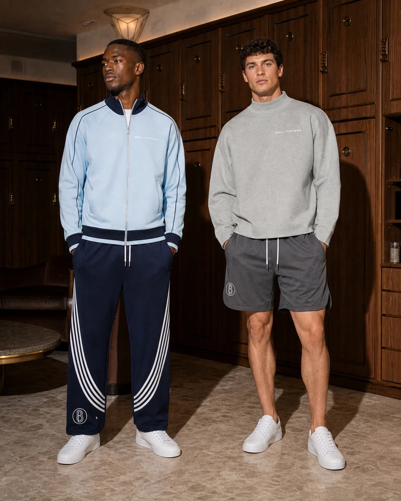 Two men model blue trackwear and a gray sweatshirt with performance shorts in a private-club locker lounge.