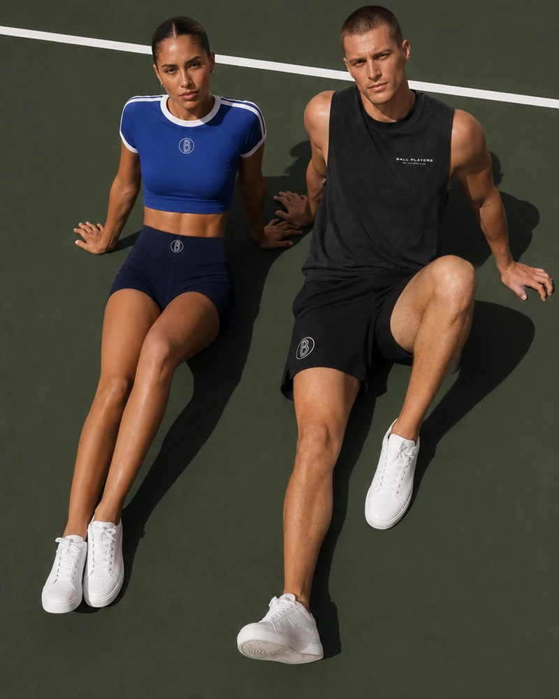 A woman and man model a blue crop top and black tank on a green tennis court.