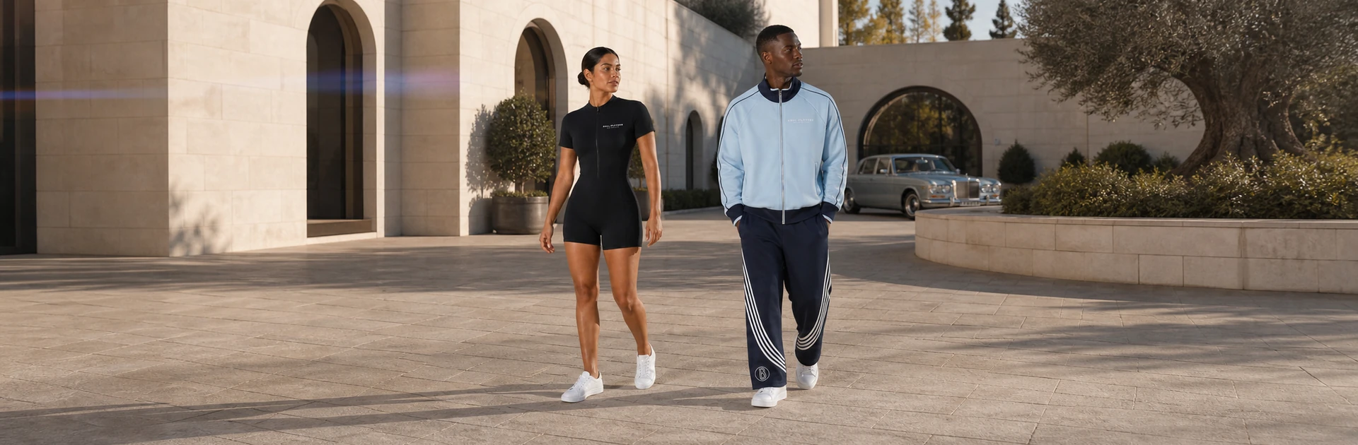 A woman in a black Ball Players romper and a man in blue trackwear cross a luxury private-club motor court.